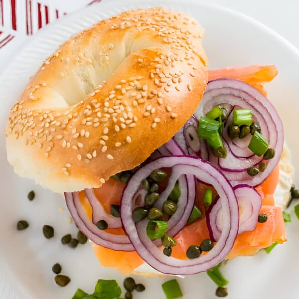 A sesame bagel with lox, red onion, capers, and green onions on a white plate.